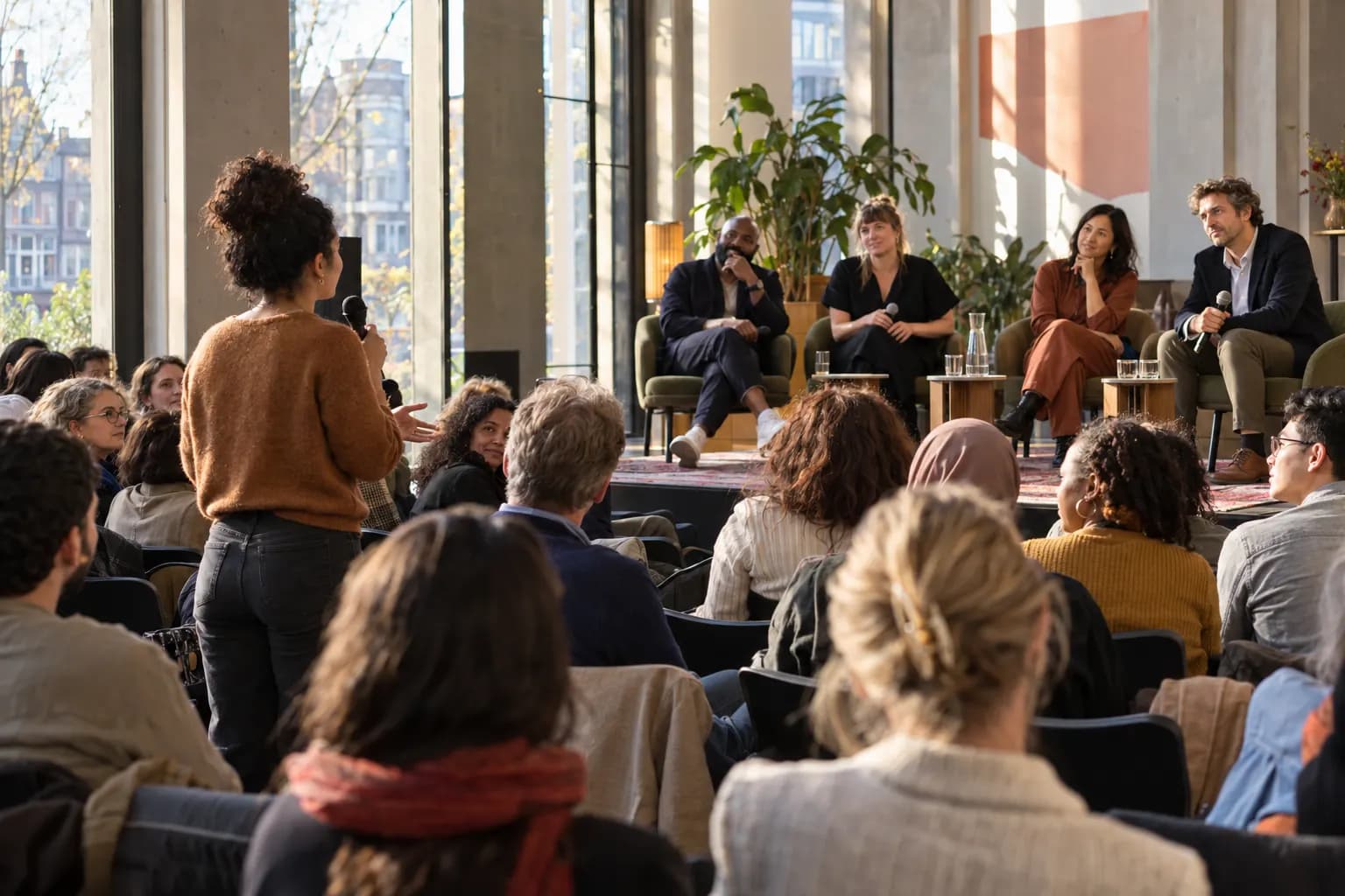 A woman in the audience asks a question at a panel discussion in a light-filled cultural centre, while four panelists listen